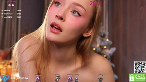 Media: A close-up video of a young, fair-skinned woman with long blonde hair and light makeup, wearing a festive, off-the-shoulder top, in a cozy, dimly-lit room with a Christmas tree.