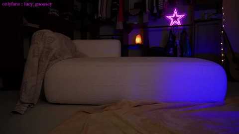 Media: A video of a dimly-lit room featuring a beige chaise lounge with a blanket draped over it. In the background, a purple star-shaped light glows, and various items are scattered on shelves.