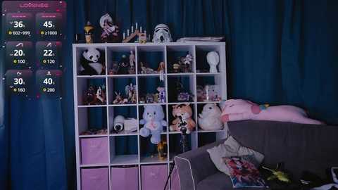 Media: Video of a cluttered room with a white shelving unit filled with stuffed toys, plushies, and a pink plush bunny on a grey couch. A digital screen displays a live stream with a chat.