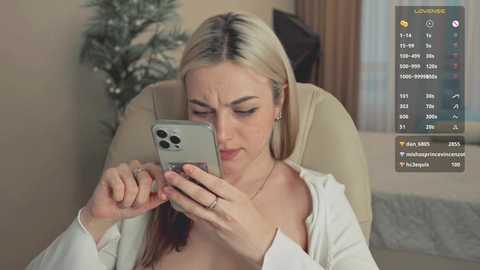 Media: Video of a blonde woman with medium skin tone, wearing a white cardigan, looking at her phone. Background shows a beige chair, a potted plant, and a window with curtains.