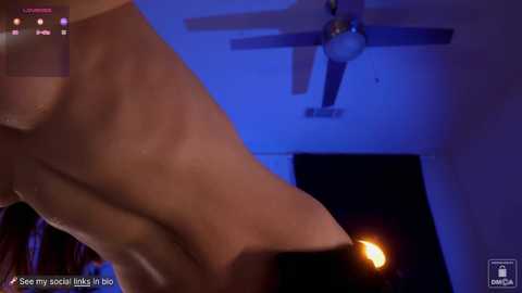 Media: A video with a low-angle view of a nude woman's foot and leg, partially illuminated by a blue light. A ceiling fan with multiple blades is visible in the background, and the room appears dimly lit.