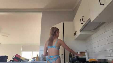 Media: Video of a blonde woman in light blue lingerie, adjusting pots on a stove in a modern kitchen with white cabinets and subway tiles.