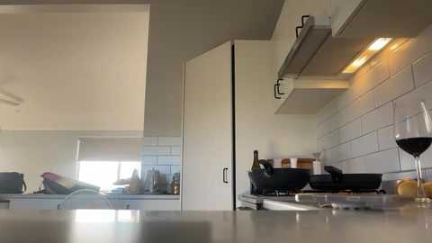 Media: A video of a modern, minimalist kitchen with white cabinetry, a stainless steel sink, white tiled backsplash, and a glass of red wine on the countertop.