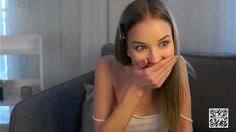 Media: Video of a young Caucasian woman with long blonde hair, wearing a white tank top, covering her mouth with her hands, sitting on a gray couch in a modern living room.