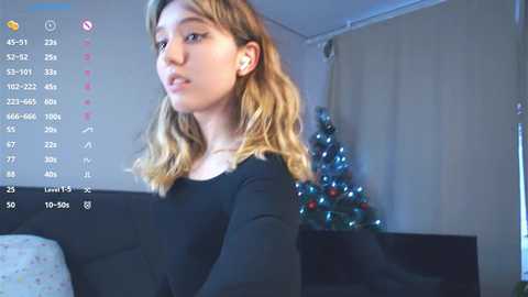 Media: A video of a young Caucasian woman with wavy, shoulder-length blonde hair, wearing a black top, standing indoors near a decorated Christmas tree.