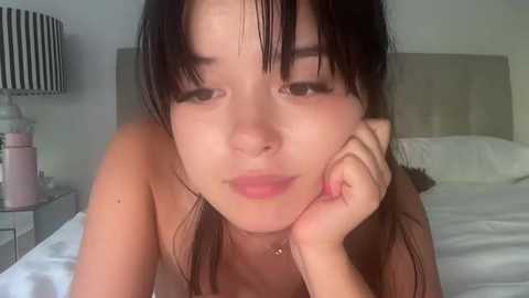 Media: A young Asian woman with long, straight black hair and fair skin, lying on a bed, topless, with a relaxed expression, resting her chin on her hand.