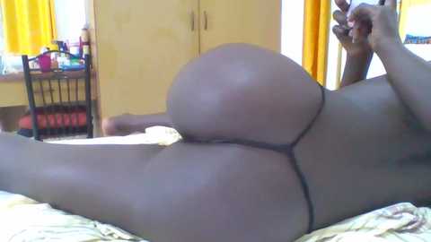 Media: Video of a naked Black woman lying on her side on a bed, wearing only a black thong. Her large, round buttocks are prominently displayed. The room features yellow curtains, a wooden dresser, and a black chair.