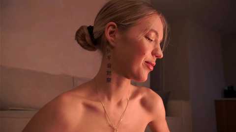 Media: Video of a topless, fair-skinned woman with blonde hair in a bun, wearing a dainty necklace. She has a tattoo with Japanese characters on her neck. Soft lighting, minimal background.