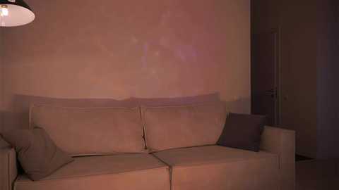 Media: A video of a dimly lit living room with beige sofa, gray cushions, and a textured, off-white wall; a black lampshade hangs overhead.