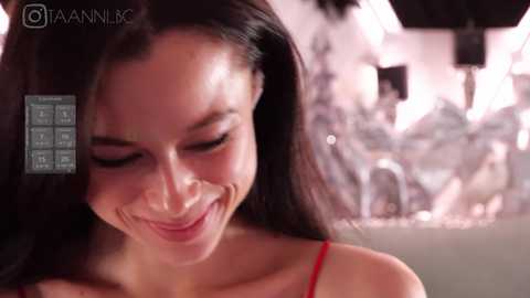 Media: Video of a smiling woman with long dark hair, wearing a red dress, in a dimly lit room with blurred, white furniture in the background.