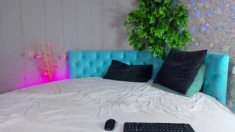 Media: Video of a modern bedroom with a turquoise tufted headboard, white bedding, green pillows, and a lush green wall-mounted plant. Pink LED lights accent the wall, creating a vibrant and cozy ambiance.