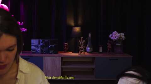 Media: Video of a dimly lit, modern room with a woman in the foreground, a wooden table in the background with a lamp, vase, and framed photo.