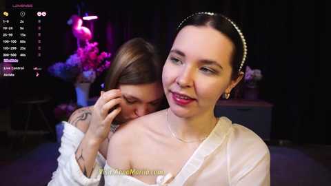 Media: Video of two women in a dimly lit room; one, with a pearl headband, wearing a white off-shoulder dress, smiles; the other, brunette with a tattoo, leans in affectionately.