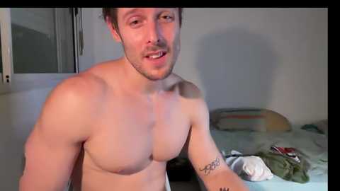 Media: Video of a shirtless, muscular, fair-skinned man with short brown hair and a trimmed beard, standing in a dimly lit room with a bed, pillow, and green clothes.