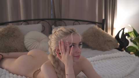 Media: A video of a nude, fair-skinned blonde woman with braided hair, lying on a bed with white sheets, surrounded by pillows and curtains, smiling while covering her eyes.