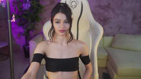 Media: Video of a young, slender woman with fair skin, black hair in a ponytail, wearing a black off-shoulder crop top and black shorts, seated on a white, ornate gaming chair in a dimly lit room with purple lighting and a potted plant.