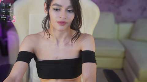 Media: Video of a slim, light-skinned, androgynous person with dark hair, wearing a black off-the-shoulder crop top, sitting in a modern, light-colored armchair in a dimly-lit room with purple walls.