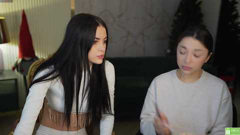 Media: Video of two women with long black hair, one in a white crop top with fringe, the other in a white sweater, seated in a modern living room with Christmas decorations and a dark green couch.