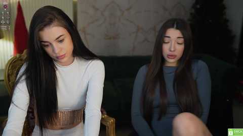 Media: Video of two young women with long, straight black hair, wearing white and blue long-sleeve tops, seated indoors. One is focused, the other relaxed. Background includes a dark green couch and Christmas tree.