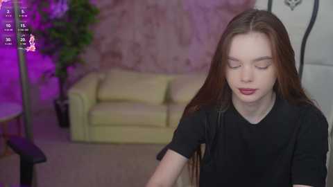 Media: Video of a young woman with long, straight brown hair, fair skin, and full lips, wearing a black top, standing in a room with a beige couch, green plant, and purple lighting.