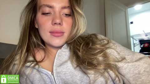 Media: Video of a young, fair-skinned woman with long, wavy blonde hair, wearing a light grey zip-up hoodie, puckering her lips in a playful manner. Background shows a white door and a cluttered room.