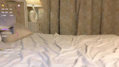 Media: Video of a cozy, beige-patterned bed with a fluffy white blanket, a bedside table holding a lamp, and soft, textured curtains in the background.