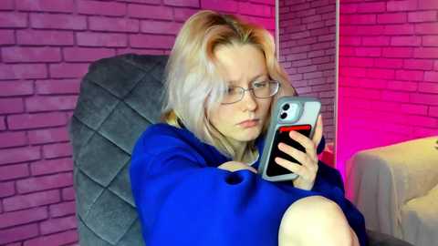 Media: Video of a blonde woman with glasses, wearing a blue sweater, seated on a gray armchair, holding a smartphone, against a brick wall with pink lighting.
