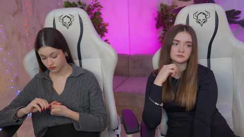 Media: Video of two young women, one with long hair in a grey sweater, the other with long hair in a black top, sitting in white gaming chairs with skull logos, under purple lighting.