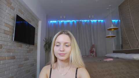 Media: Video of a blonde woman in a modern bedroom with a stone wall, TV, blue LED lighting, and hanging pendant lights.