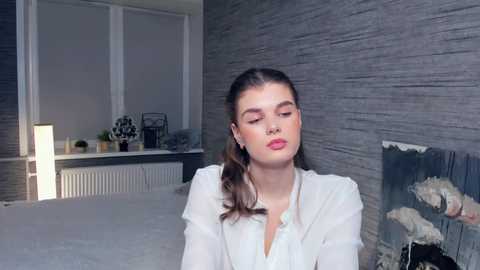 Media: Video of a young woman with fair skin, brown hair, and light makeup, wearing a white blouse, standing in a modern, dimly lit room with gray textured walls, white radiator, and abstract art on the right.