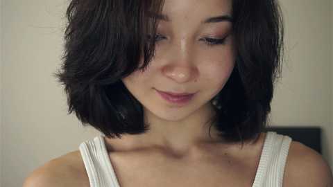 Media: Video of a young woman with short, dark hair and light skin, wearing a white tank top, smiling softly with closed eyes. Background is a blurred beige wall.