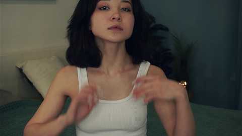 Media: A video of a young woman with long black hair, fair skin, and slender build, wearing a white tank top, adjusting her bra straps in a dimly lit room with a beige wall and green carpet.