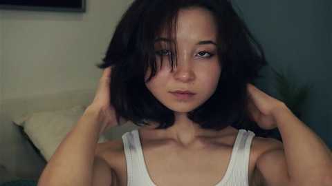 Media: A video of a young Asian woman with short, dark hair, wearing a white tank top, adjusting her hair in a dimly lit room.