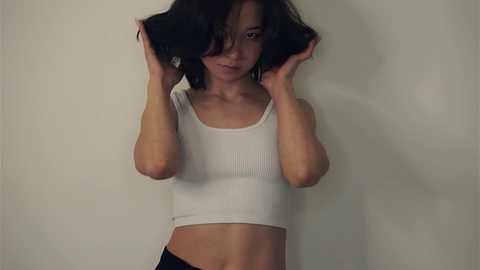 Media: Video of a young Asian woman with medium-length dark hair, wearing a white ribbed tank top and black shorts, standing against a plain, light-colored wall, playing with her hair.