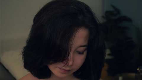 Media: Video of a young girl with shoulder-length, dark brown hair partially covering her face, smiling softly, standing indoors against a blurred background of green foliage and dim lighting.