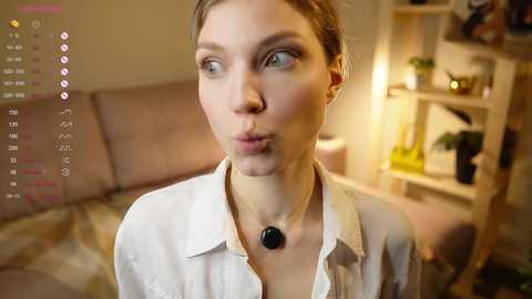 Media: Video of a young woman with fair skin, light brown hair, and blue eyes, wearing a white button-up shirt with a black pendant necklace, pouting playfully. Background shows a beige couch and a wooden shelf with plants.