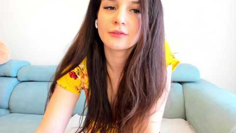 Media: Video of a young woman with long, straight, dark brown hair, fair skin, and a slender physique, wearing a yellow floral top, seated on a light blue couch against a plain white wall.