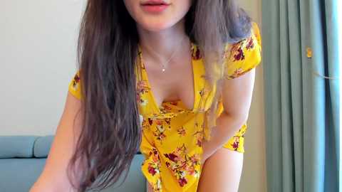 Media: Video of a woman with long, straight brown hair, wearing a yellow floral dress with a deep V-neck, leaning forward, indoors with light blue curtains and a light-colored couch in the background.