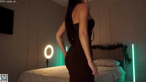 Media: Video of a slender woman with long, dark hair, wearing a tight, strapless brown dress, standing in a dimly lit bedroom with a bed, a round light, and a Christmas wreath.