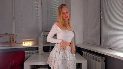 Media: Video of a blonde Caucasian woman with long hair, wearing a white off-shoulder crop top and a light blue ruffled skirt, standing in a modern, brightly lit room with white walls and windows.