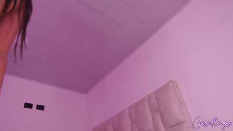 Media: Video of a woman's face and shoulders, partially visible, in a room with pink lighting, beige tufted headboard, and white ceiling.