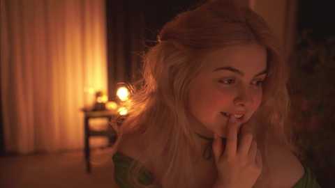 Media: Video of a blonde woman with fair skin, wearing a green dress, sitting in a dimly-lit room with a warm, yellow light. She has a playful expression, covering her mouth with her hand.