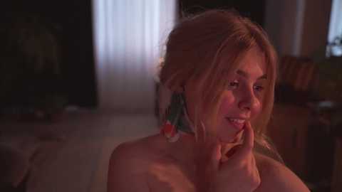 Media: Video of a young woman with light skin, blonde hair, and freckles, smiling and touching her face. She wears colorful earrings. The background is dimly lit with a window and furniture, giving a cozy, intimate atmosphere.