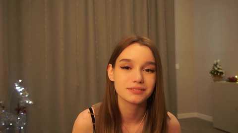 Media: Video of a young Caucasian woman with long, straight brown hair, wearing a sleeveless top, standing in a dimly lit room with gray curtains and a small, lit Christmas tree in the background.