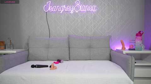 Media: Video of a modern bedroom with a gray tufted headboard, white bedding, and a neon sign reading \"Jeniffer Banca.\" The bed features a black vibrator, a pink dildo, and a peach-colored dildo.