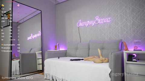 Media: A modern bedroom with a gray upholstered bed, a mirrored wall, purple lighting, and a neon sign reading \"George Barrios\" on a white textured wall.