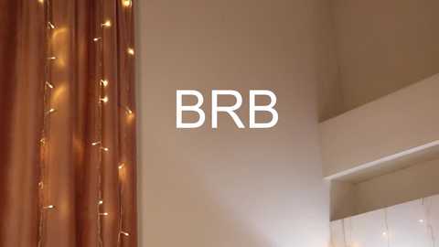 Video of a beige wall with a \"BBR\" logo and warm, golden string lights draped over a brown curtain on the left. The background features a white shelf and a soft, ambient light.