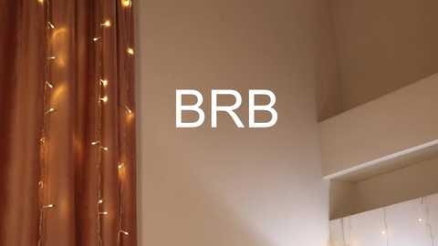 Video of a beige wall with the text \"BRB\" in white, next to a window with brown curtains adorned with warm yellow fairy lights.
