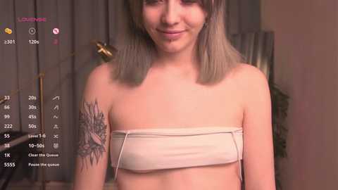 Media: Video of a smiling, light-skinned woman with shoulder-length, blonde hair, wearing a beige tube top, revealing a detailed tattoo on her left shoulder. Background shows a dimly lit room with grey curtains.