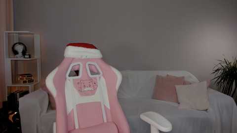 Media: Video of a pink gaming chair with a Santa hat, white couch with pink and beige pillows, beige walls, white shelves with books, and a potted plant.